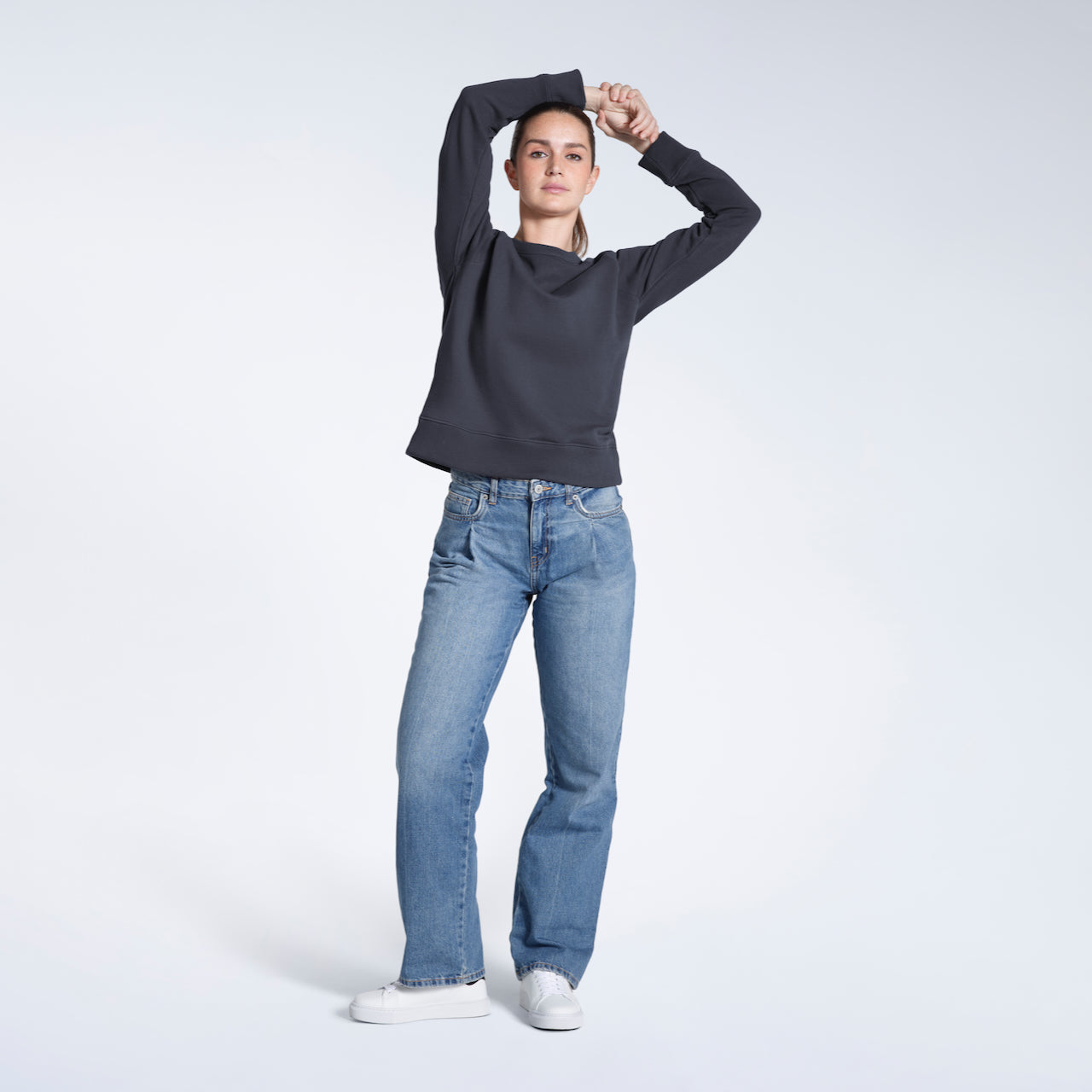 A full body photo of a woman looking into the camera with her hands above her head, modelling a plain dark grey french terry organic cotton sweatshirt.
