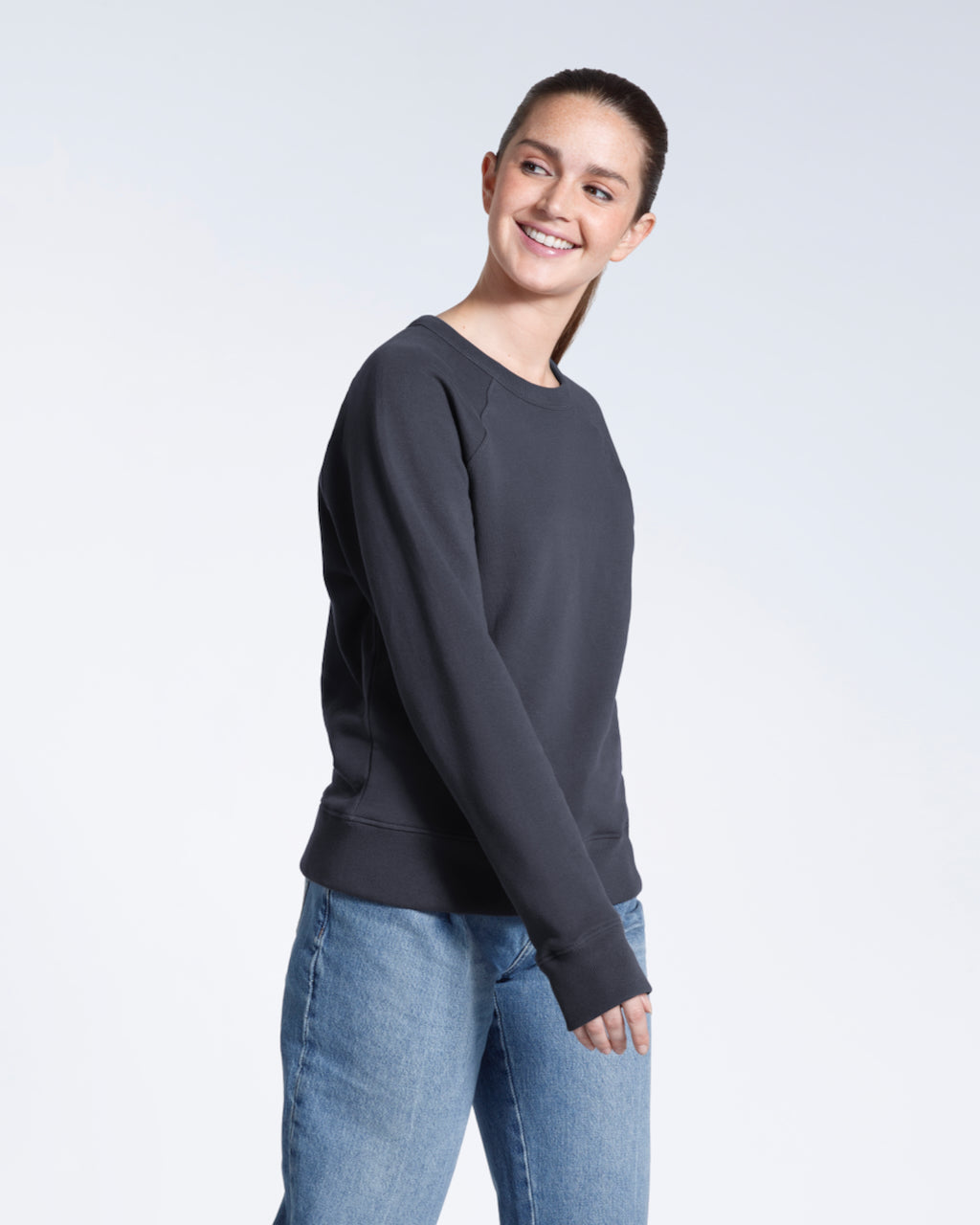 A woman smiling looking away from the camera, modelling a plain dark grey french terry organic cotton sweatshirt.
