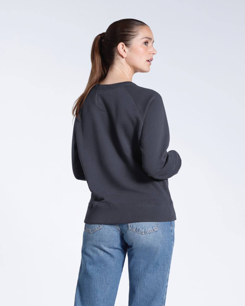 A woman facing away from the camera, showing the back of her plain dark grey french terry organic cotton sweatshirt.