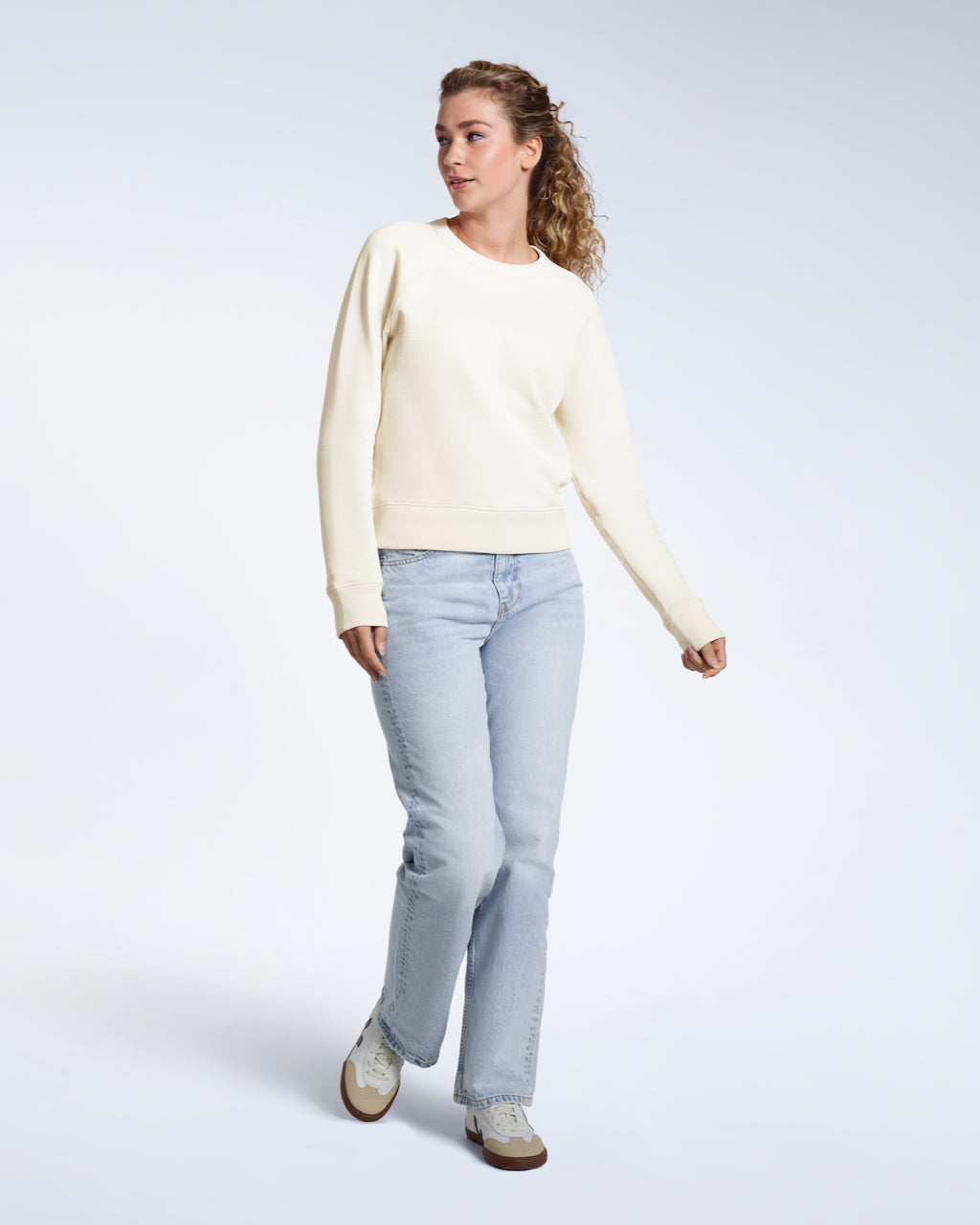 A full body photo of a woman looking away from the camera, modelling a plain ecru french terry organic cotton sweatshirt.