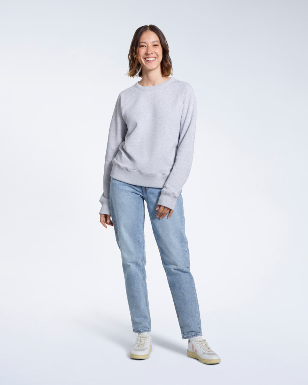 A full body photo of a woman smiling at the camera, modelling a plain grey french terry organic cotton sweatshirt.