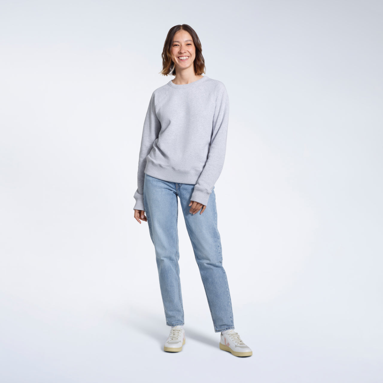 A full body photo of a woman smiling at the camera, modelling a plain grey french terry organic cotton sweatshirt.