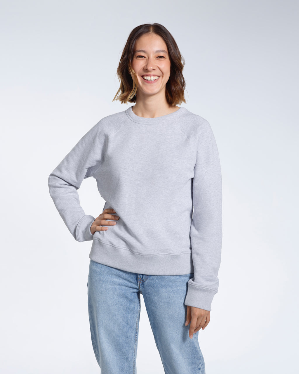A woman smiling into the camera with one hand on her hip, modelling a plain grey french terry organic cotton sweatshirt.