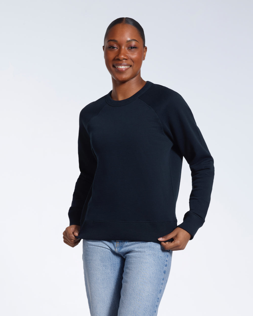 A woman smiling into the camera, touching the bottom of her plain navy french terry organic cotton sweatshirt.