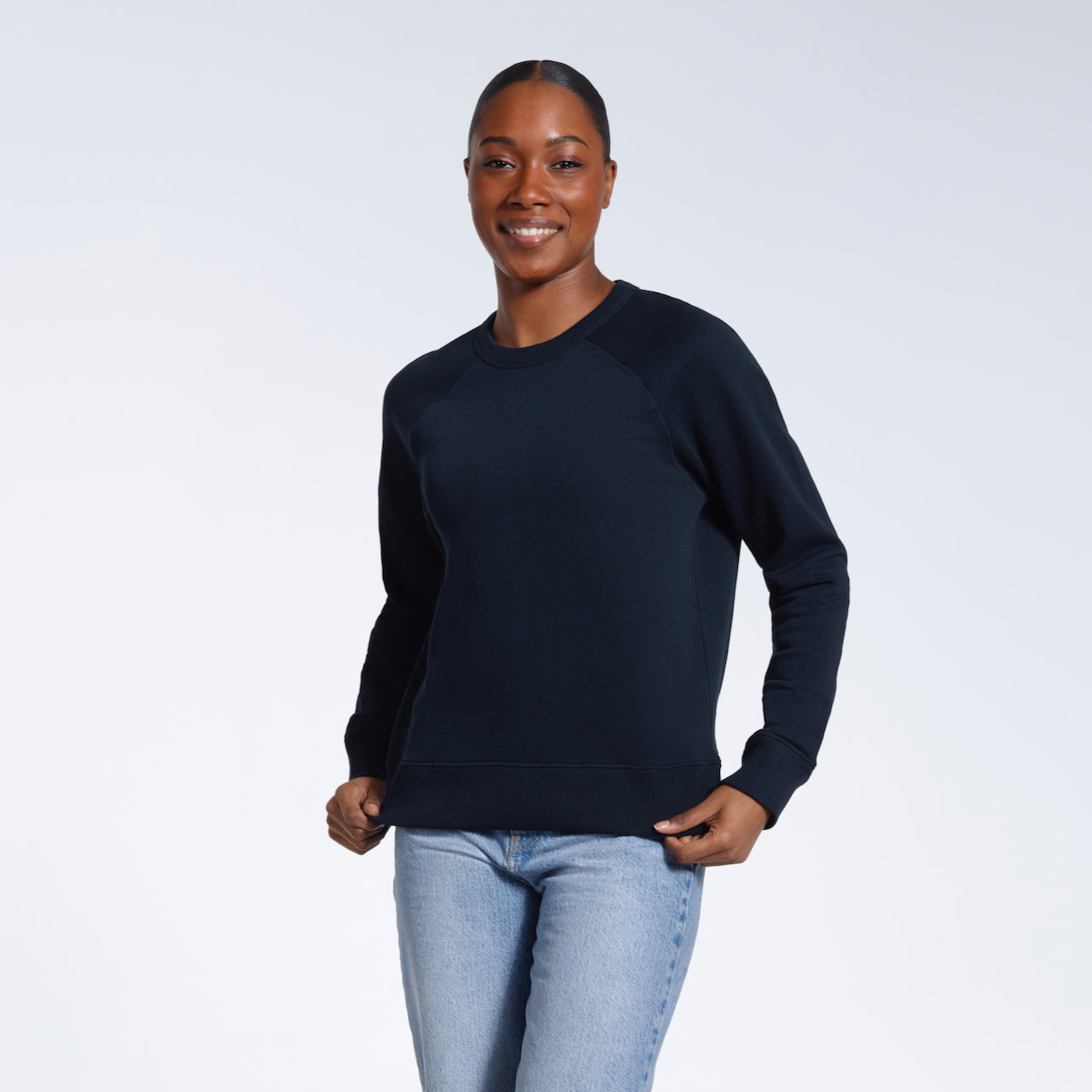 A woman smiling into the camera, touching the bottom of her plain navy french terry organic cotton sweatshirt.