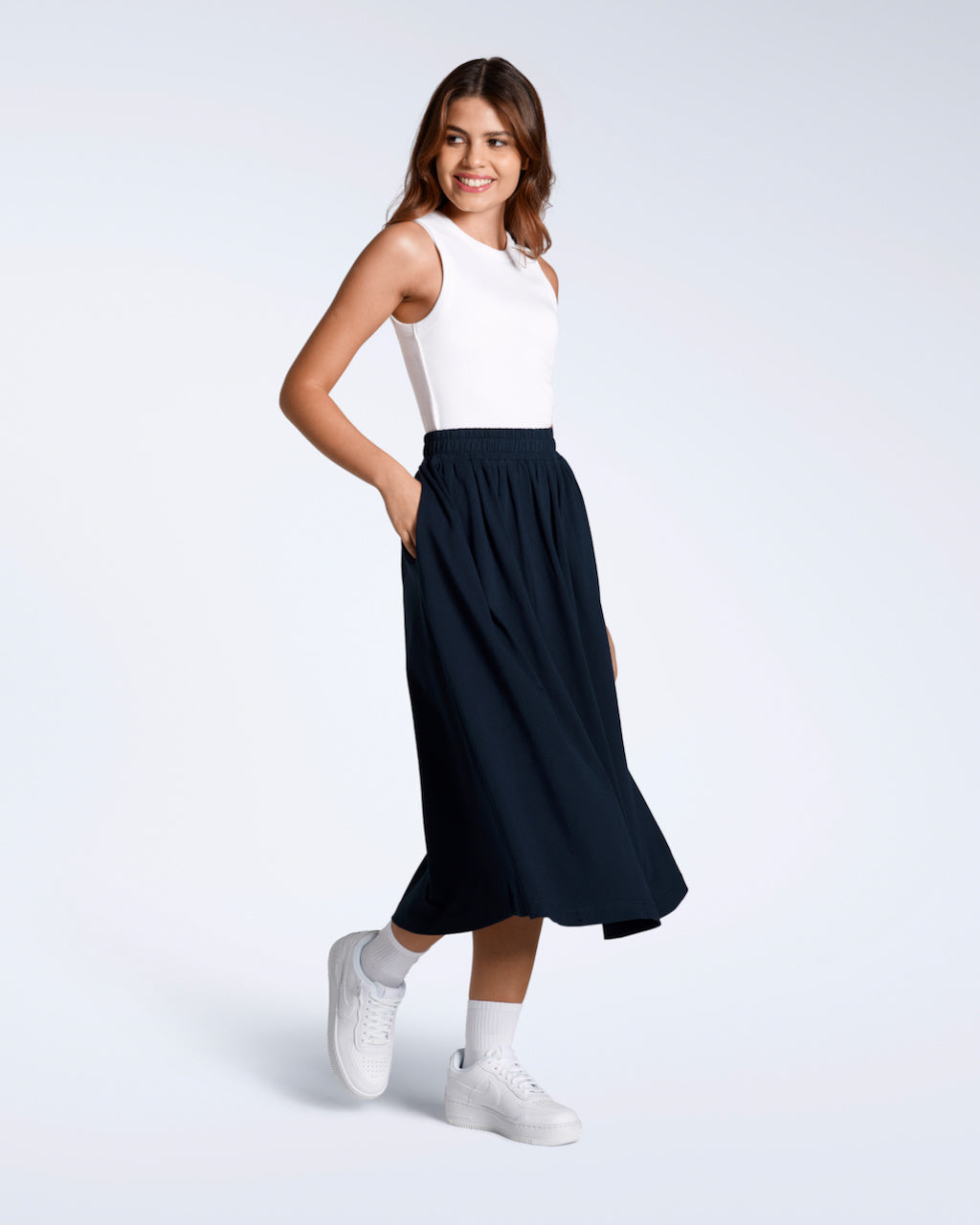 A full body photo of a woman smiling with her hand in her pocket, modelling a flowy, navy, organic cotton midi skirt.