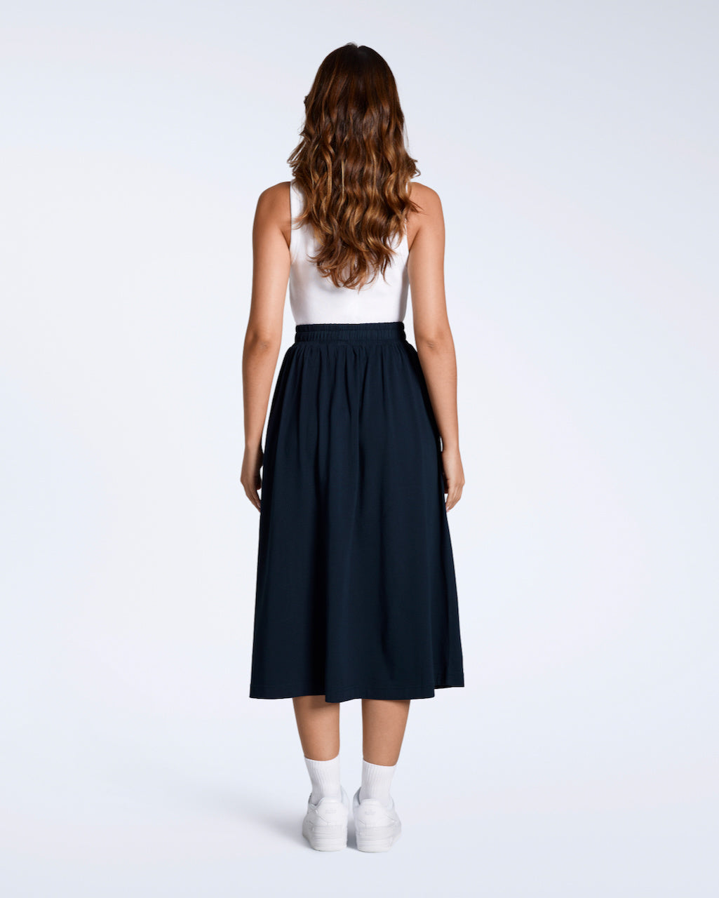 A full body photo of a woman facing away from the camera, modelling the back of a flowy, navy, organic cotton midi skirt.