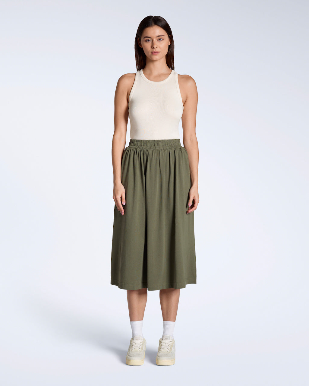 A full body photo of a woman looking into the camera with her arms by her side, modelling a flowy, olive coloured, organic cotton midi skirt.