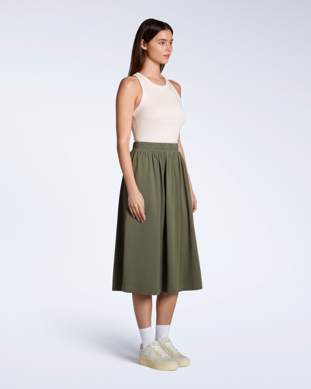 A full body photo of a woman at an angle looking away from the camera, modelling a flowy, olive coloured, organic cotton midi skirt.