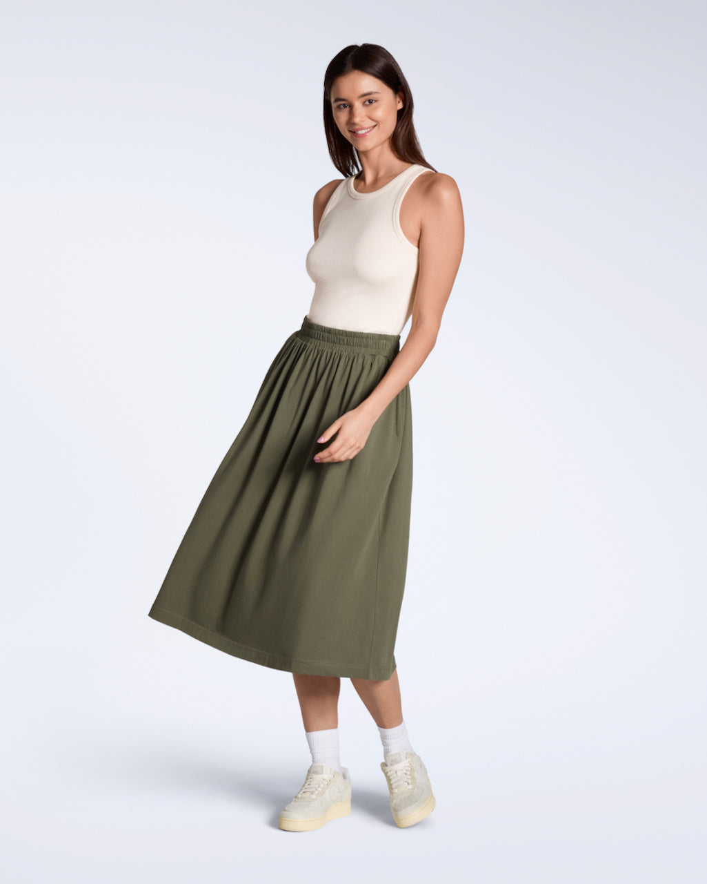 A full body photo of a woman smiling into the camera, modelling a flowy, olive coloured, organic cotton midi skirt.