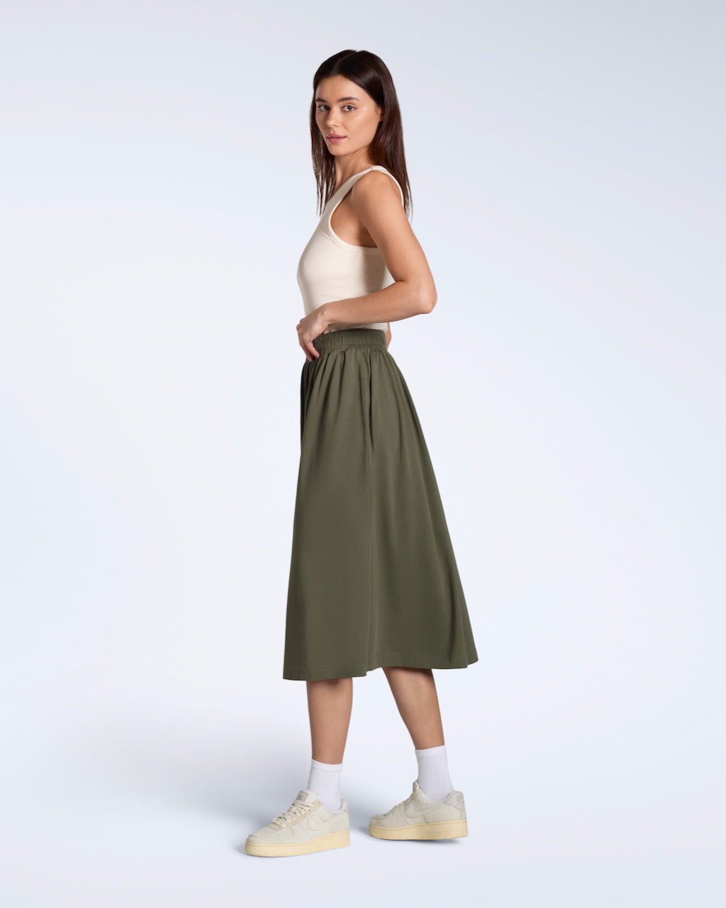 A full body photo of a woman facing side on and looking into the camera, modelling a flowy, olive coloured, organic cotton midi skirt.