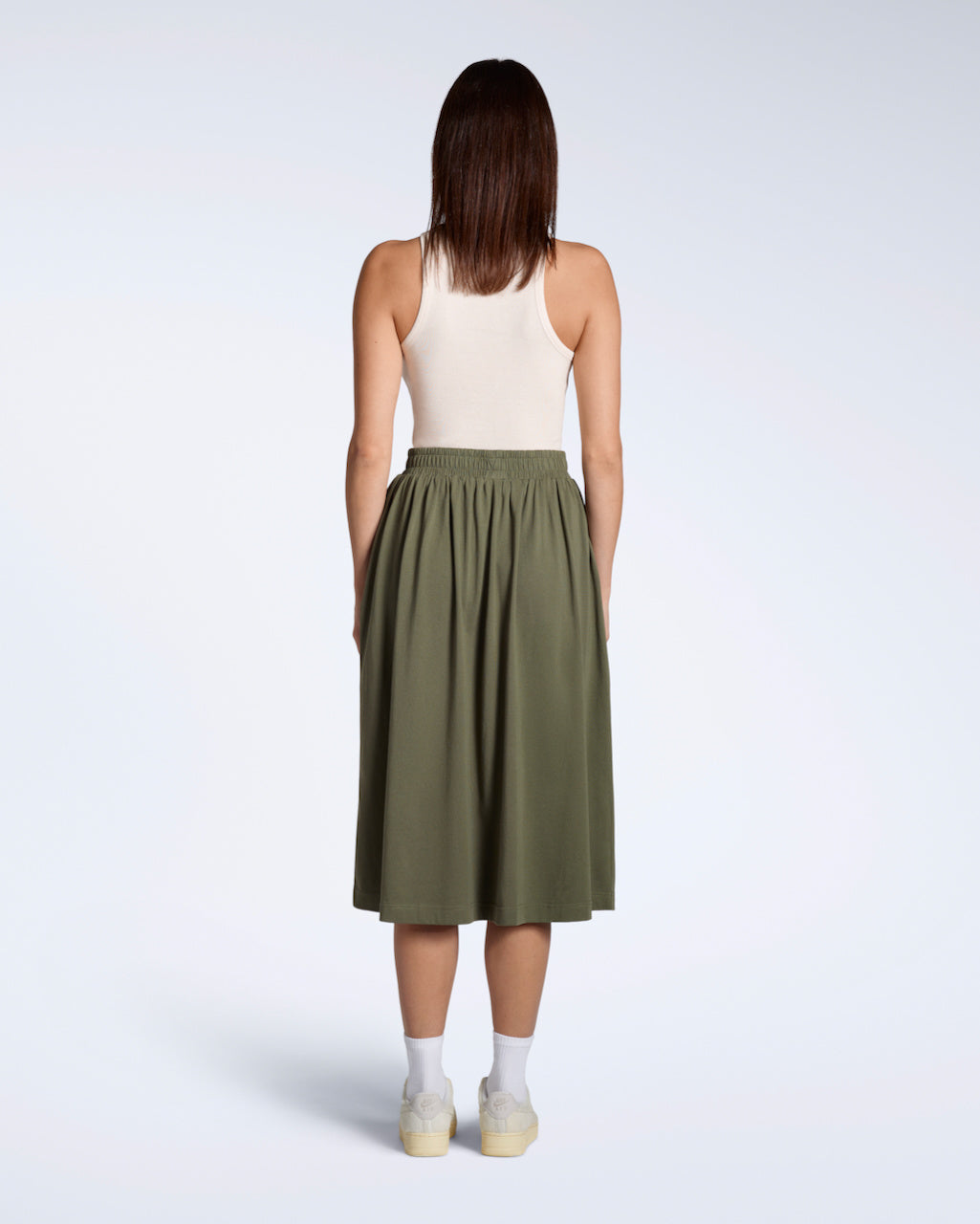A full body photo of a woman facing away from the camera, modelling the back of a flowy, olive coloured, organic cotton midi skirt.