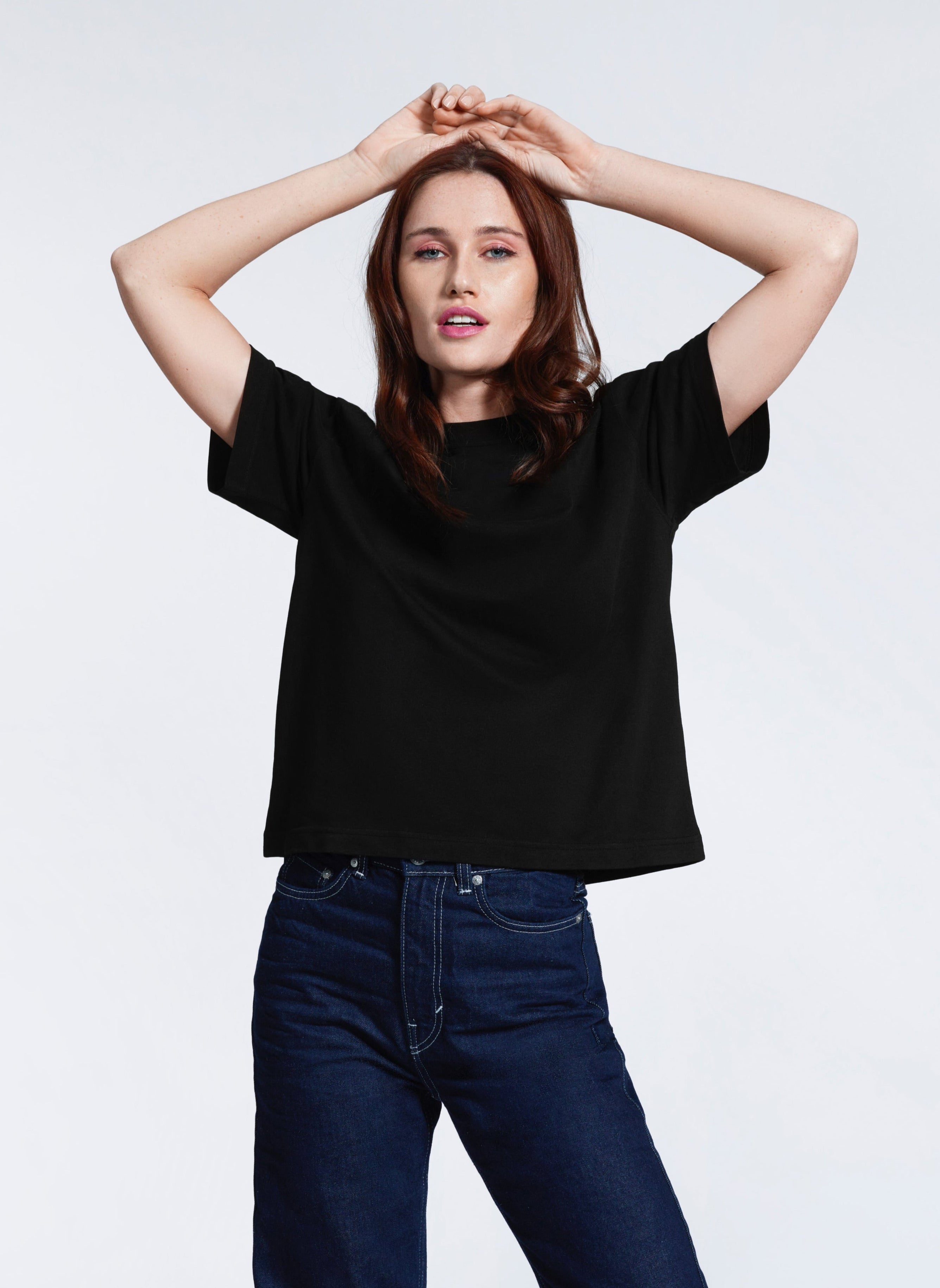 A woman with her hands on her head looking at the camera, modelling a plain black boxy, heavyweight organic cotton t shirt.