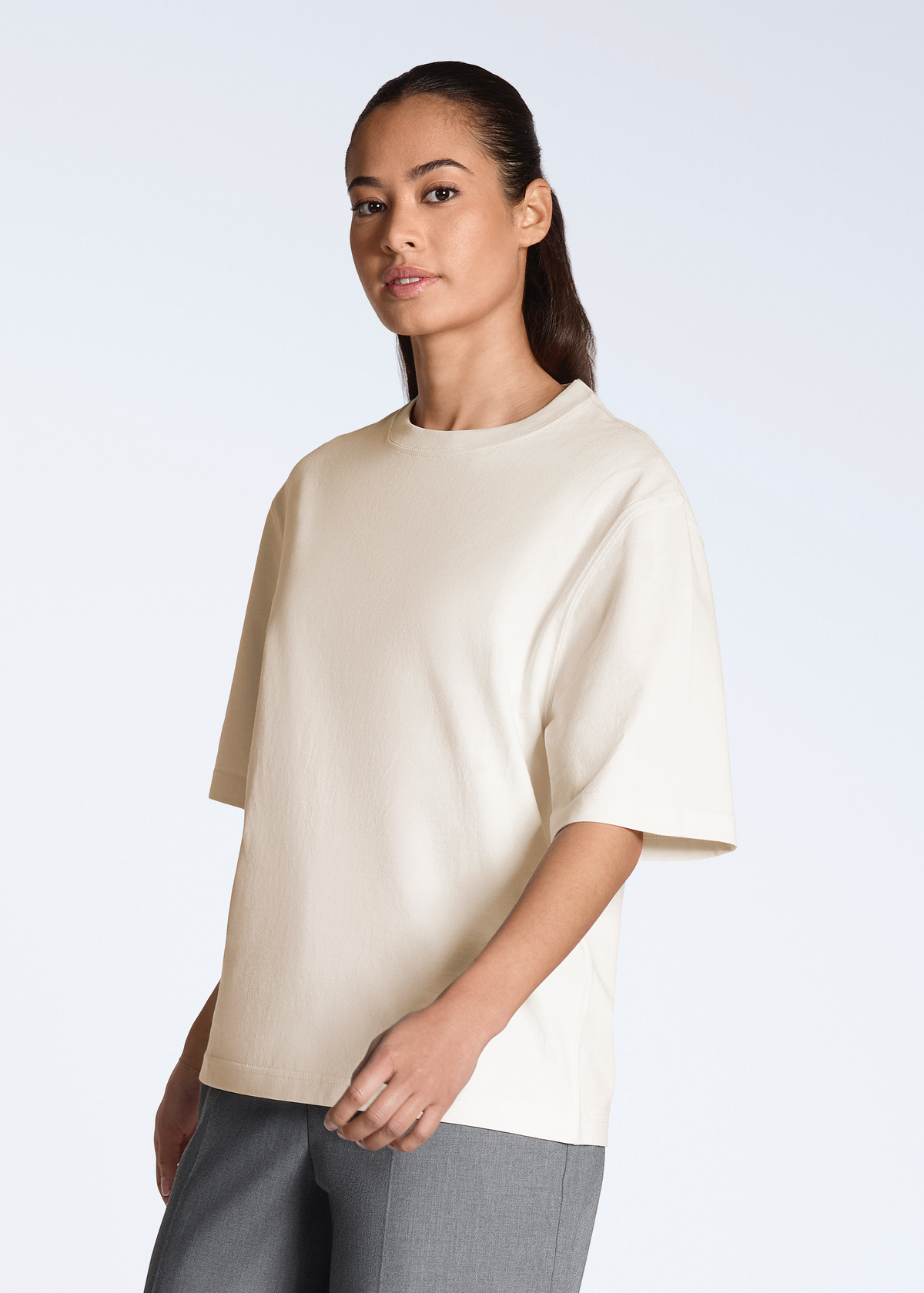 A woman looking into the camera, modelling a plain natural coloured, boxy, heavyweight, organic cotton t shirt.