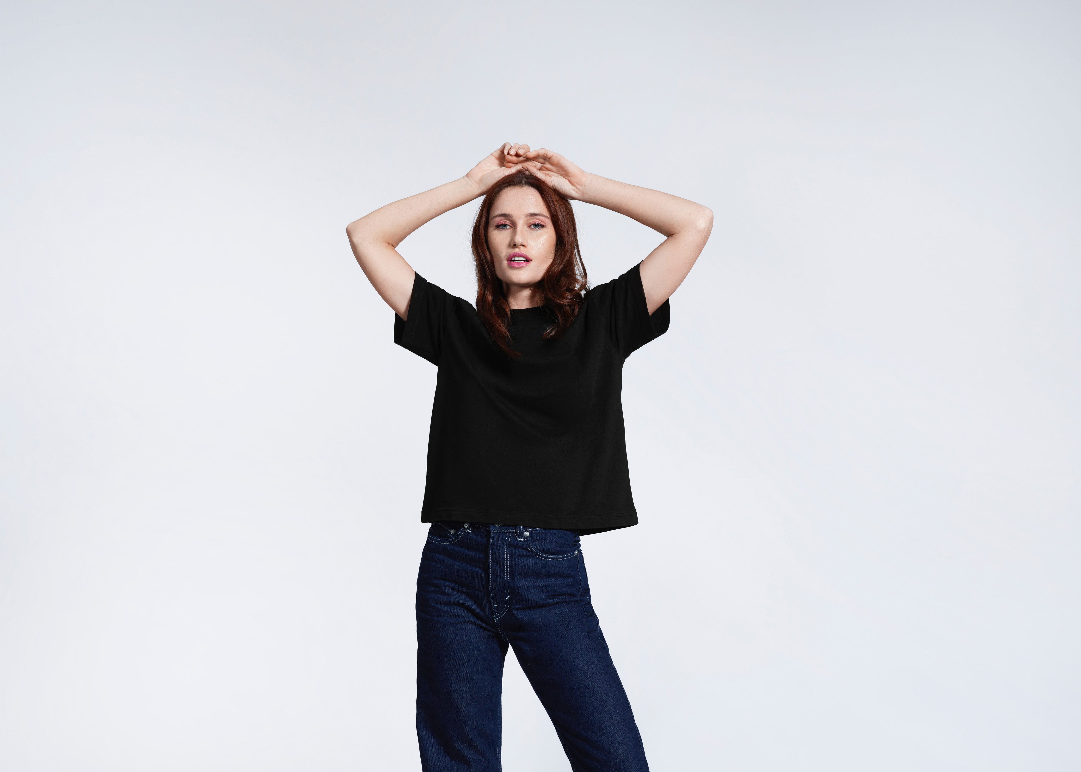 A woman with her hands on her head looking at the camera, modelling a plain black boxy, heavyweight organic cotton t shirt.