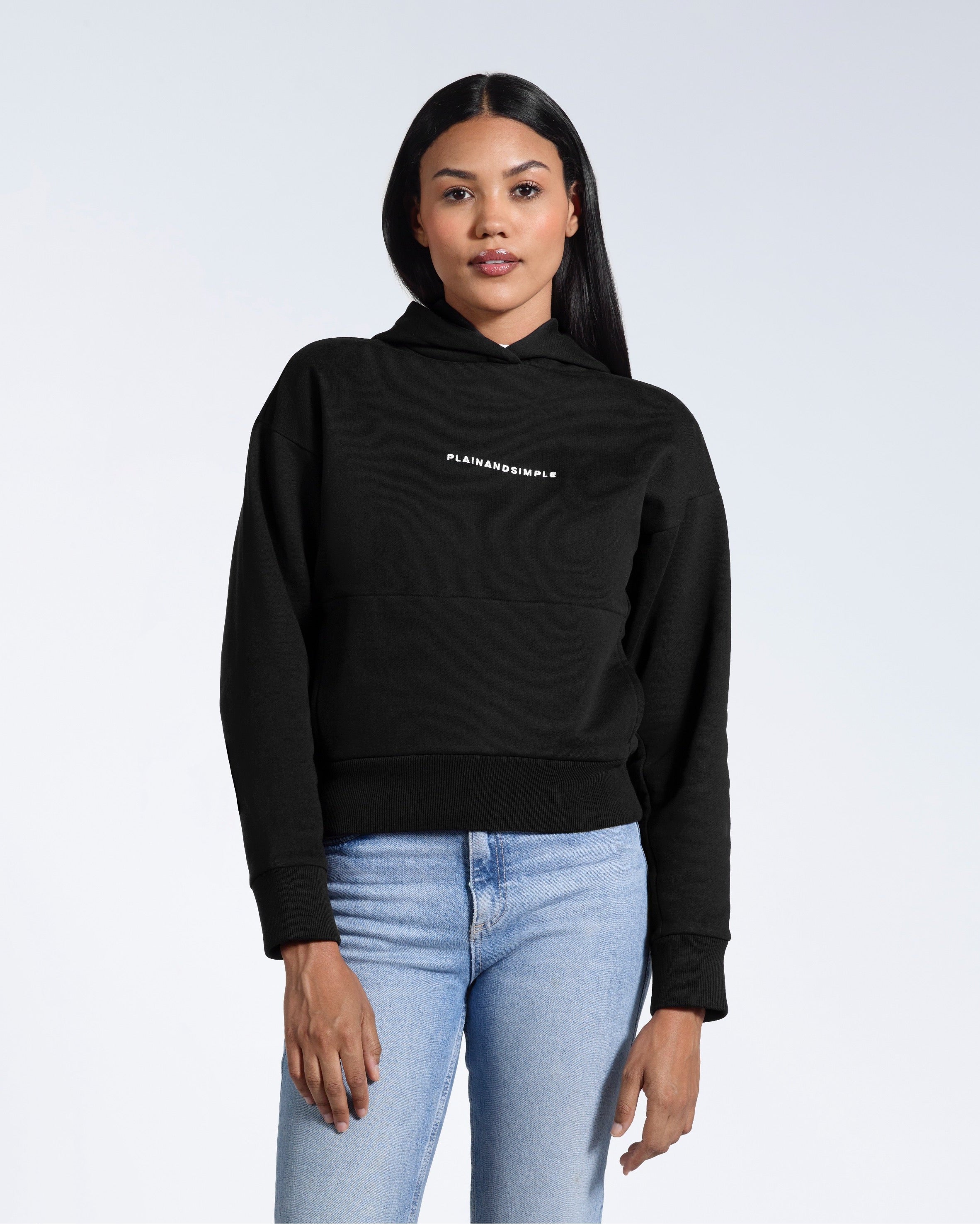 A woman facing front on looking into the camera with her arms by her sides, modelling a branded black heavyweight organic cotton hoodie.