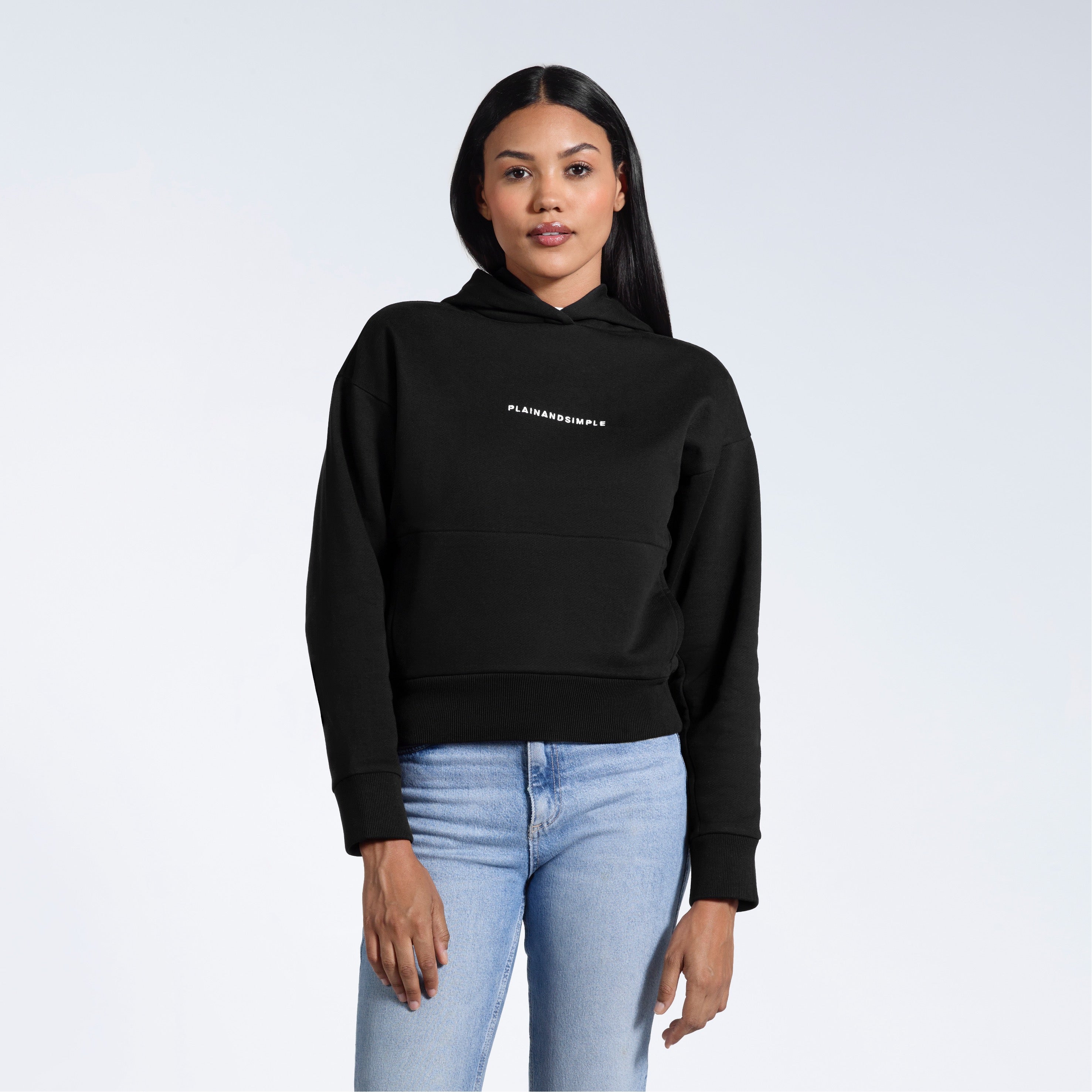 A woman facing front on looking into the camera with her arms by her sides, modelling a branded black heavyweight organic cotton hoodie.