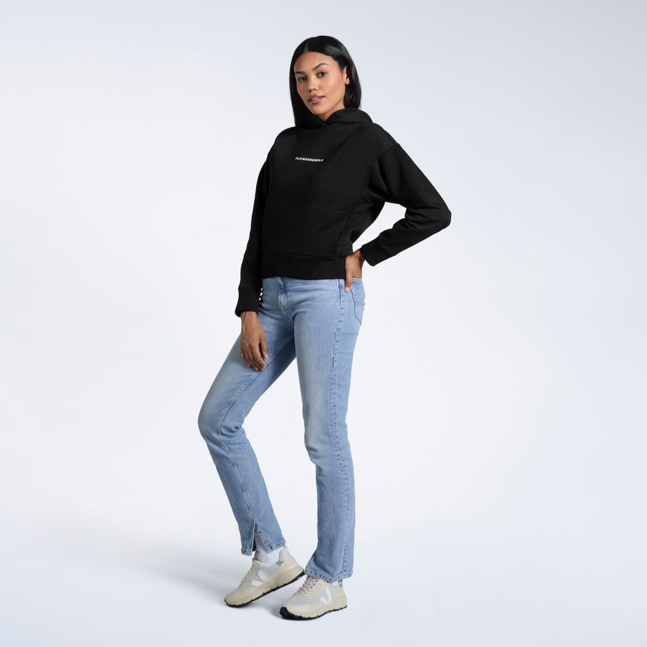 A full body photo of a woman looking into the camera with her hand in her back pocket, modelling a black branded heavyweight organic cotton hoodie.