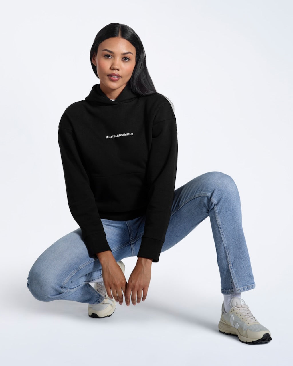 A woman crouching down, modelling a branded black heavyweight organic cotton hoodie.