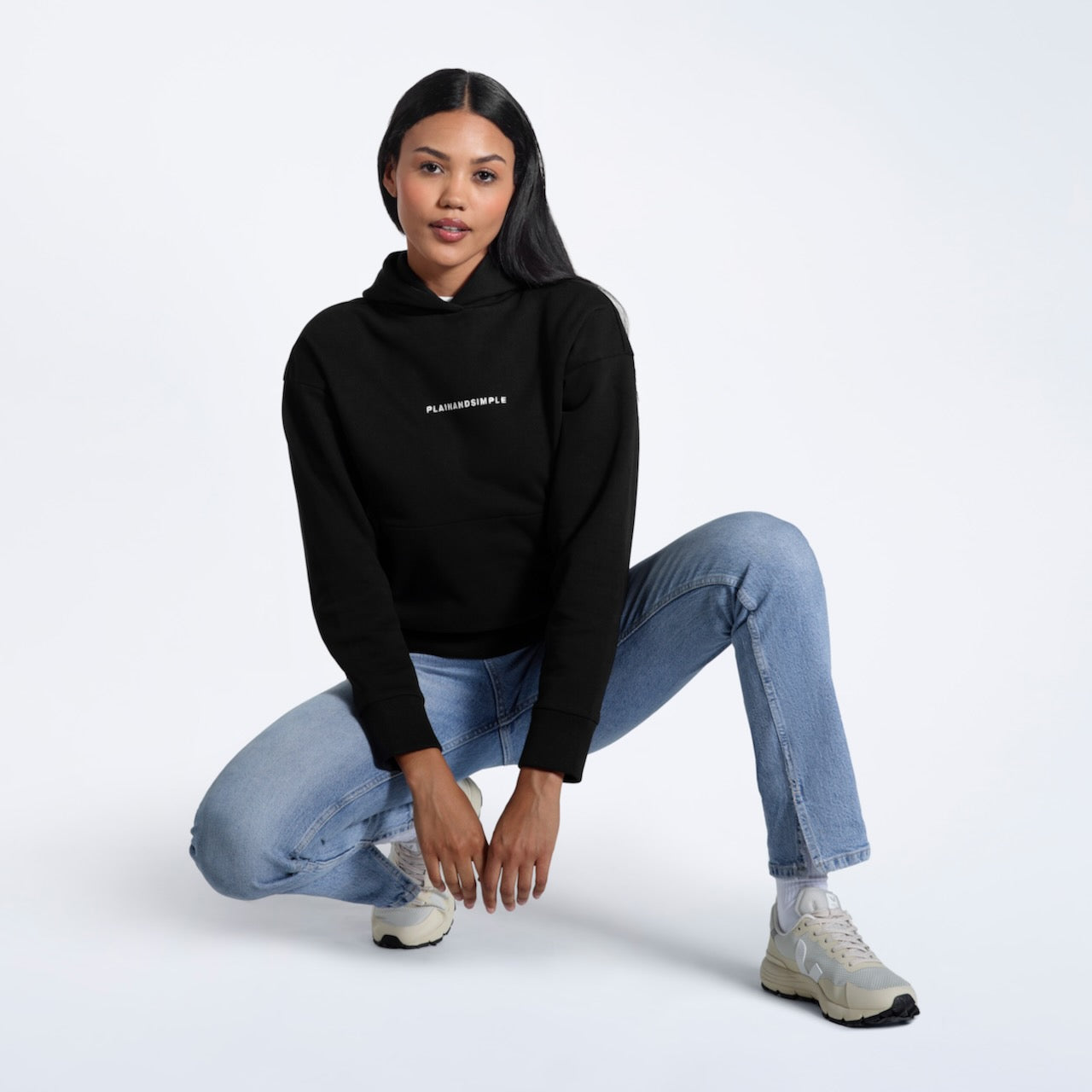 A woman crouching down, modelling a branded black heavyweight organic cotton hoodie.