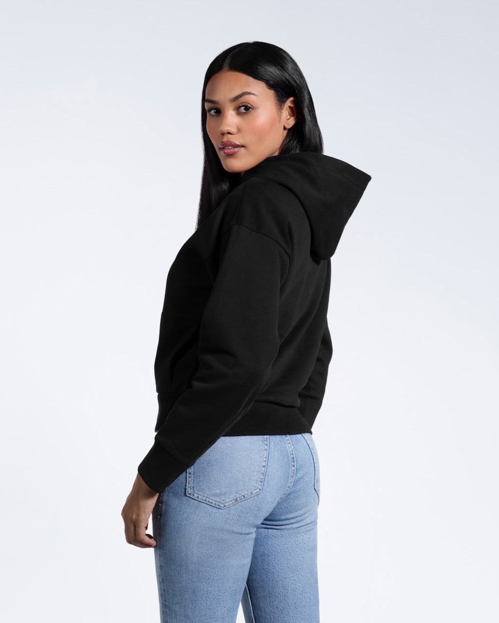 A woman with her head turned towards the camera, showing the back of her black heavyweight organic cotton hoodie.