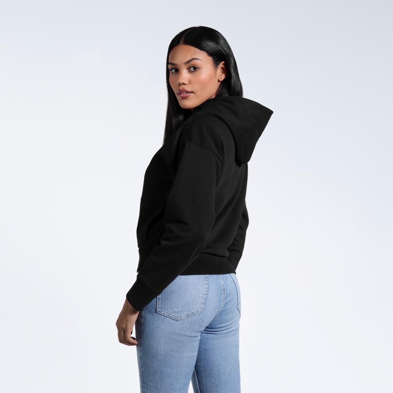 A woman with her head turned towards the camera, showing the back of her black heavyweight organic cotton hoodie.
