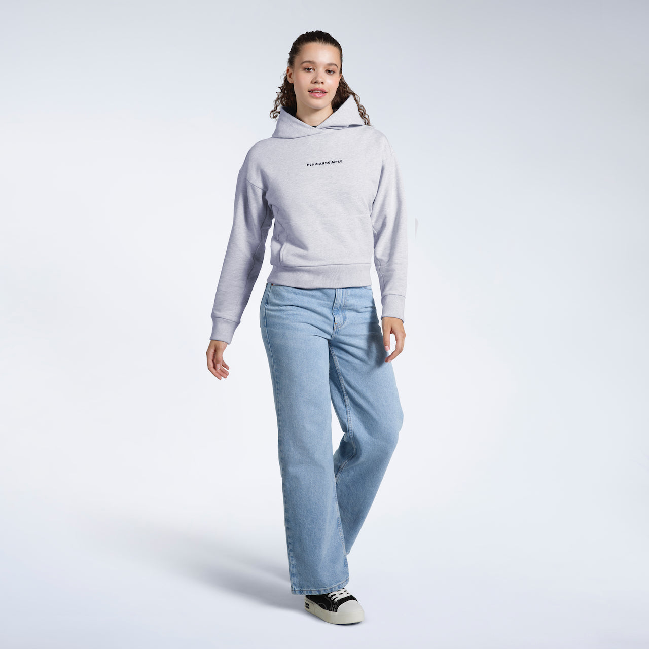 A full body photo of a woman walking looking into the camera, modelling a grey branded heavyweight organic cotton hoodie.