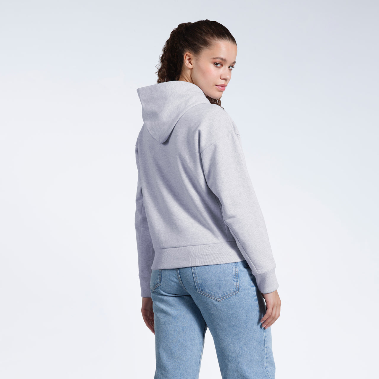 A woman with her head turned towards the camera, showing the back of her grey heavyweight organic cotton hoodie.