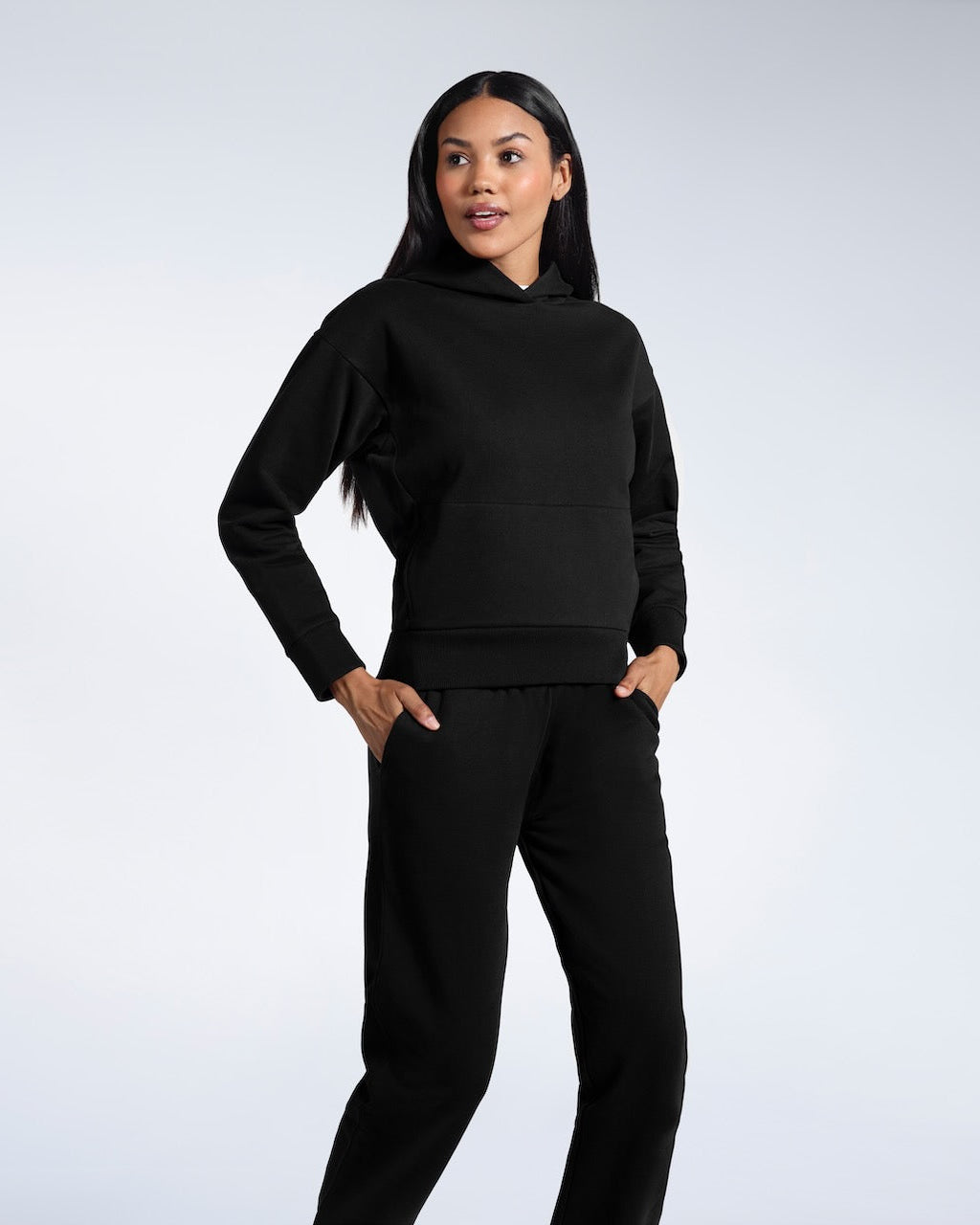 A woman with her hands in her pockets looking away from the camera, modelling a black heavyweight organic cotton hoodie with matching joggers.
