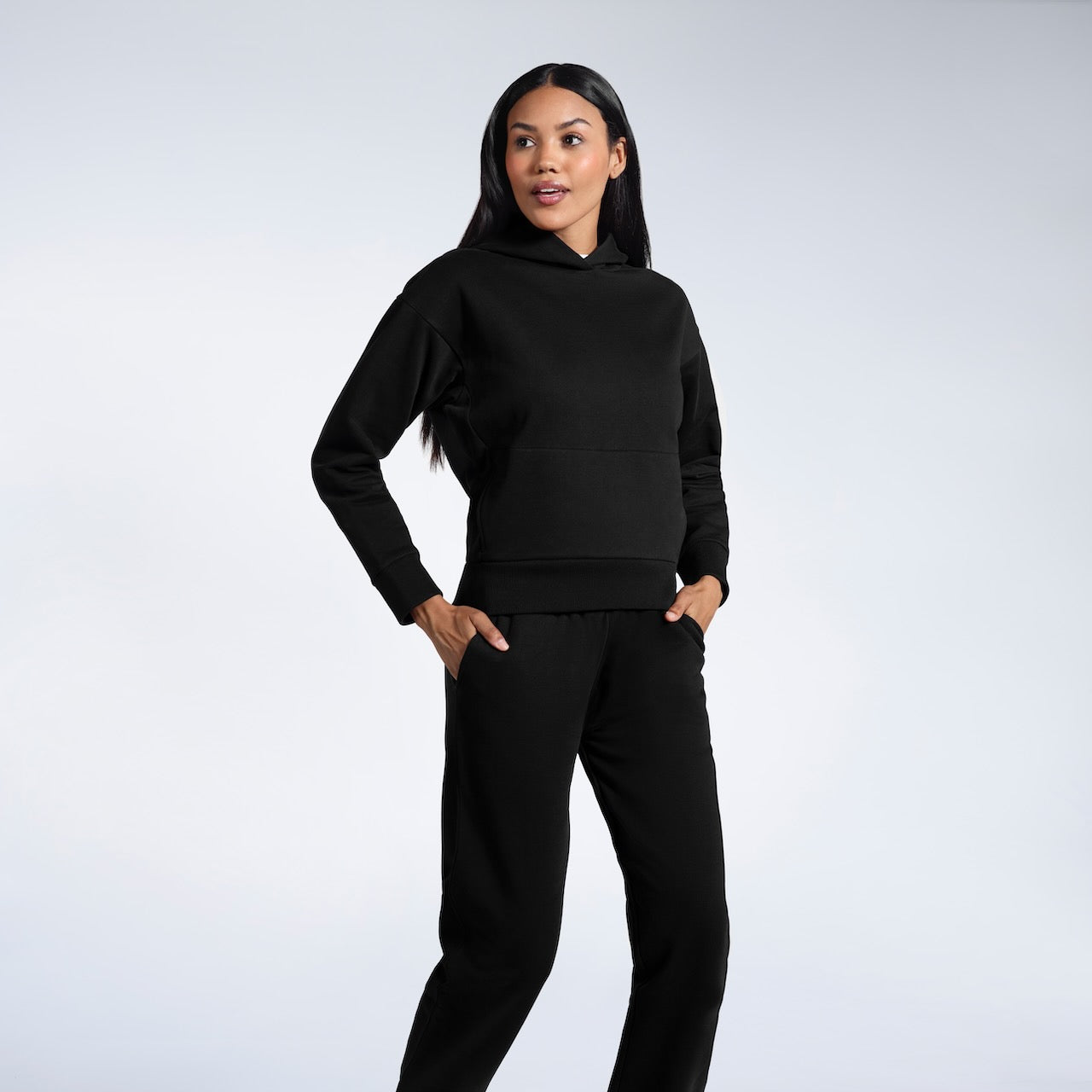 A woman with her hands in her pockets looking away from the camera, modelling a black heavyweight organic cotton hoodie with matching joggers.