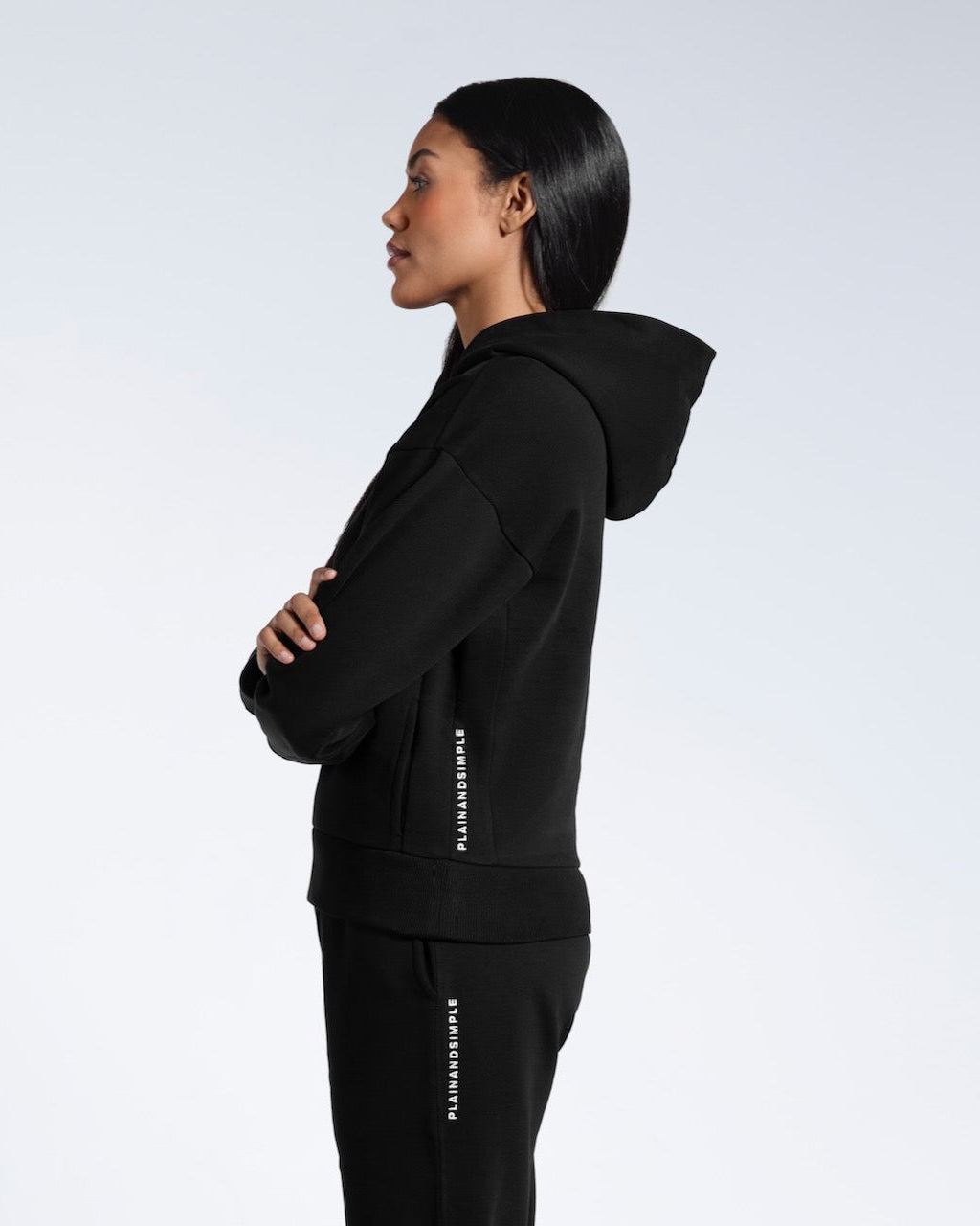 A woman facing side on, modelling a branded black heavyweight organic cotton hoodie with matching joggers.