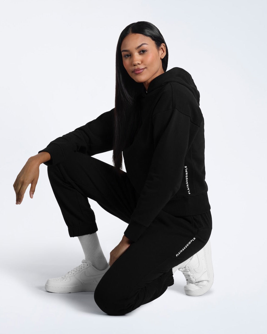 A woman crouching down, modelling a branded black heavyweight organic cotton hoodie with matching joggers.
