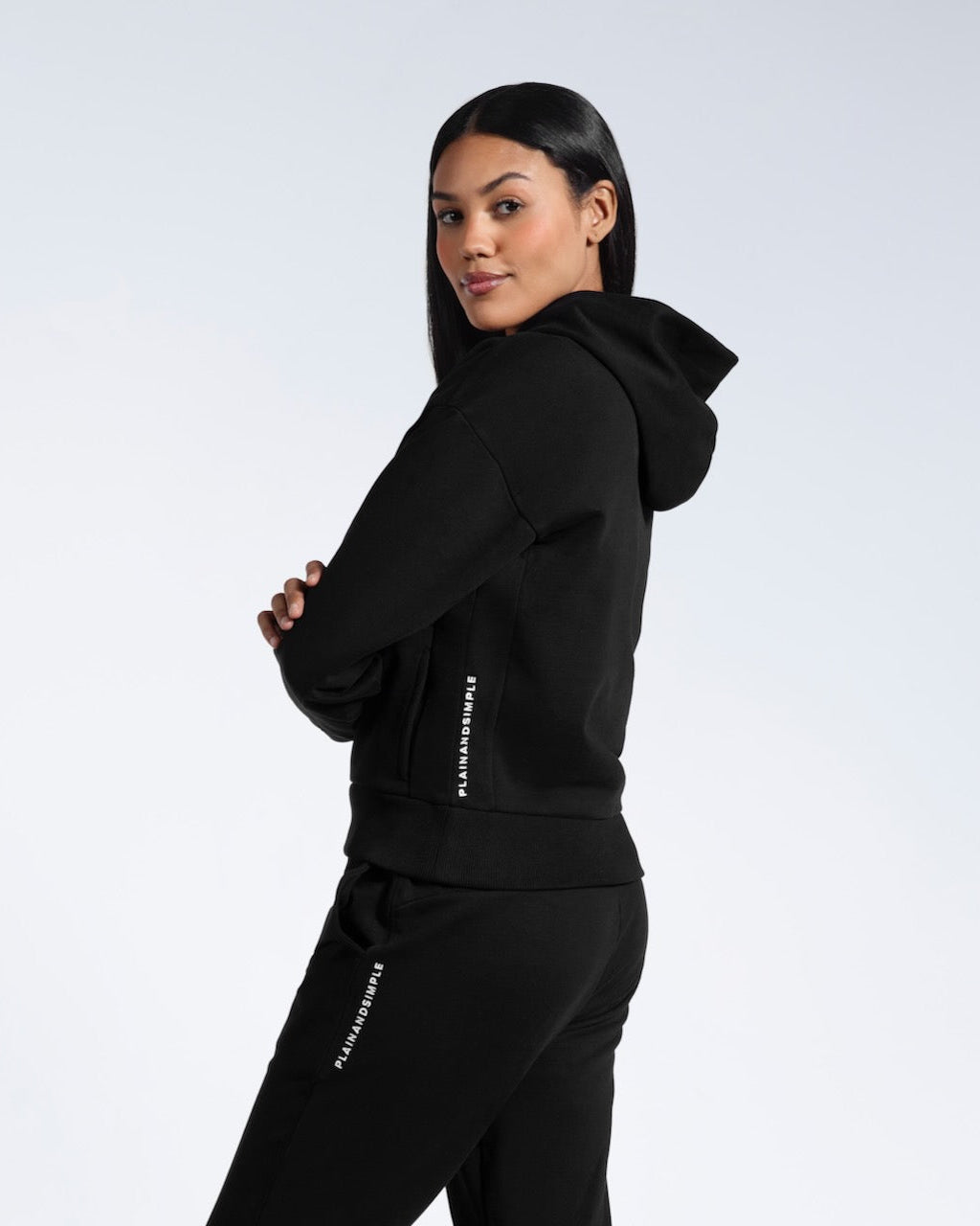 A woman facing side on looking into the camera, modelling a branded black heavyweight organic cotton hoodie with matching joggers.