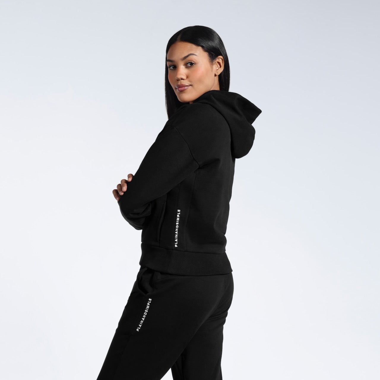 A woman facing side on looking into the camera, modelling a branded black heavyweight organic cotton hoodie with matching joggers.