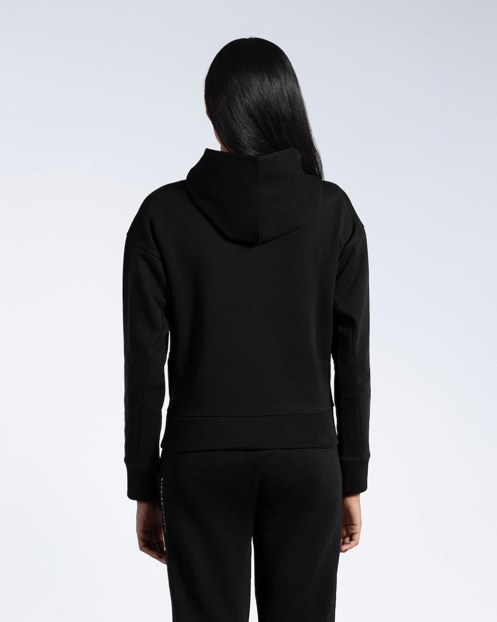A woman facing away from the camera with her arms by her sides, modelling a black heavyweight organic cotton hoodie with matching joggers.