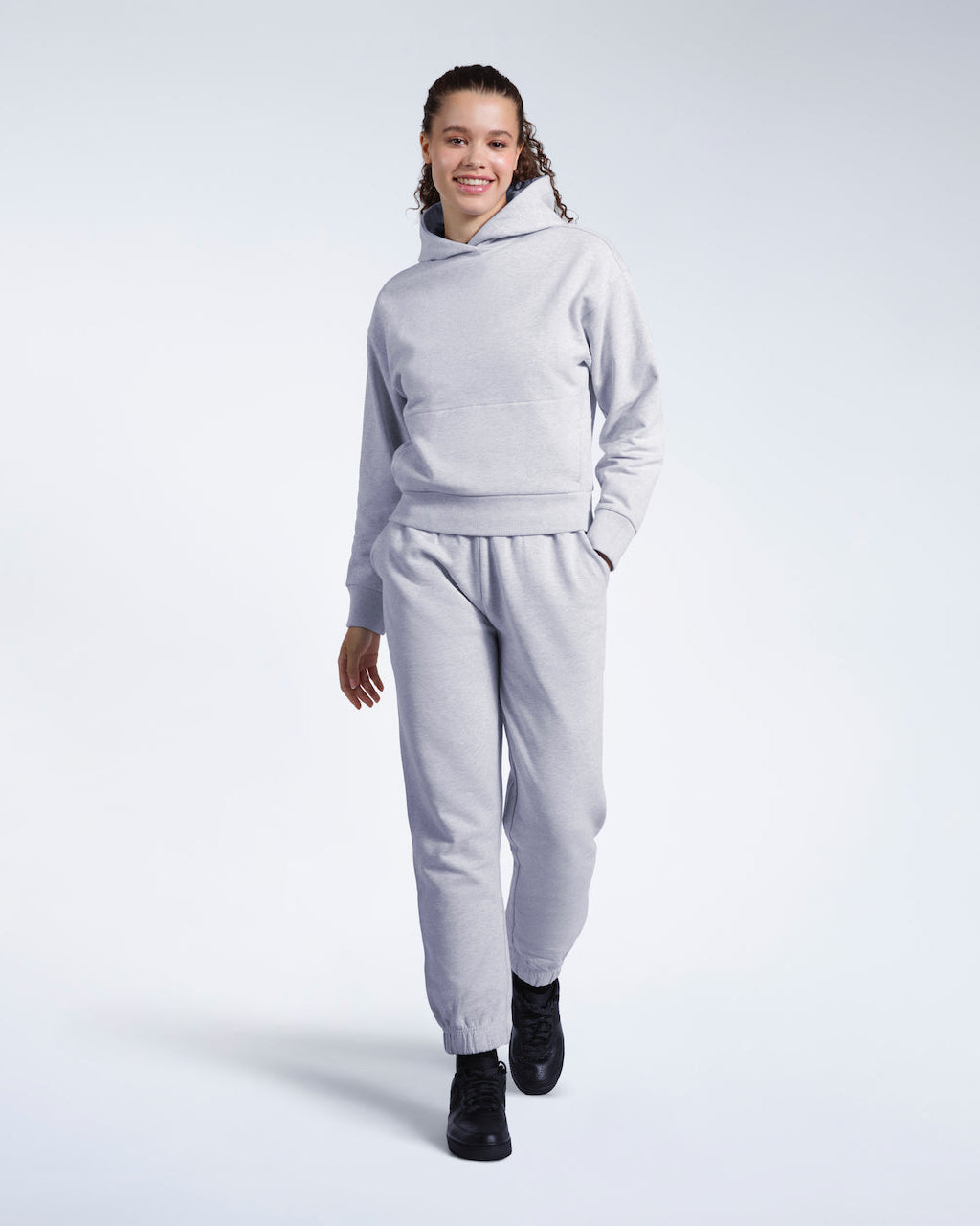 A full body photo of a woman walking and smiling into the camera, modelling a matching grey heavyweight organic cotton tracksuit.