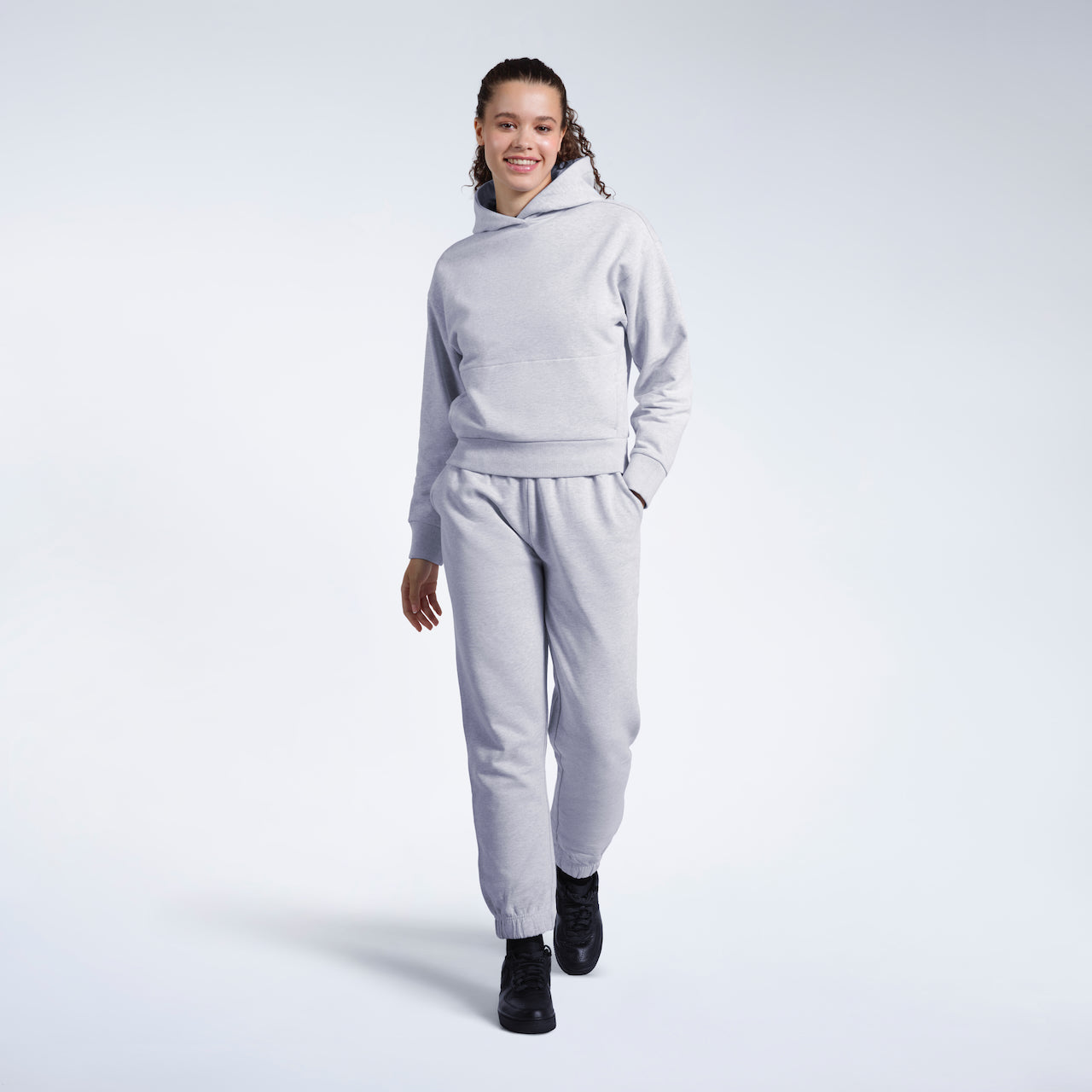 A full body photo of a woman walking and smiling into the camera, modelling a matching grey heavyweight organic cotton tracksuit.