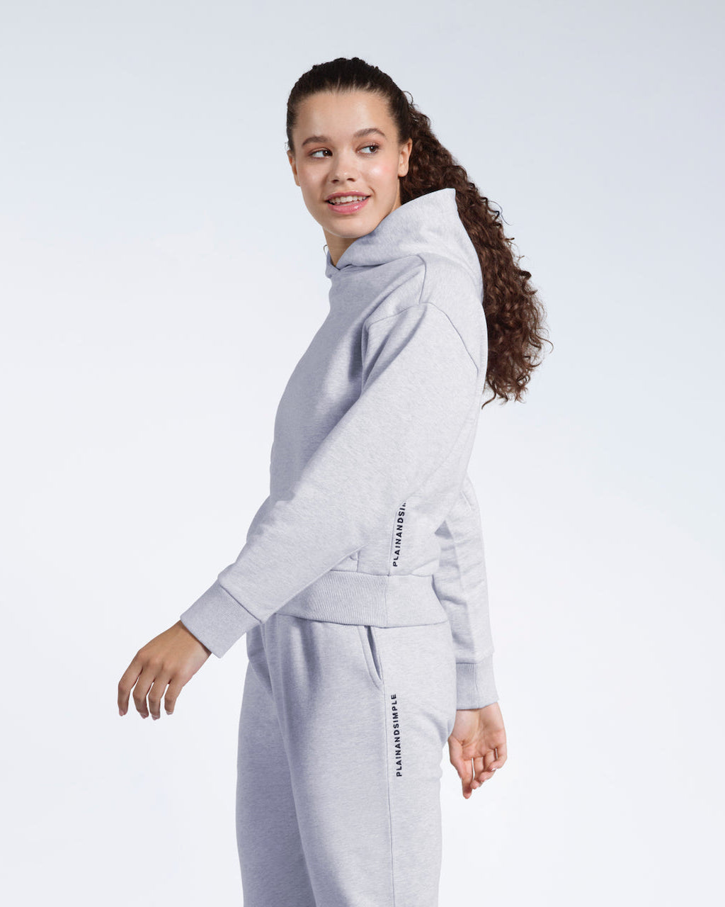 A woman mid movement looking away from the camera smiling, modelling a branded grey heavyweight organic cotton hoodie with matching joggers.