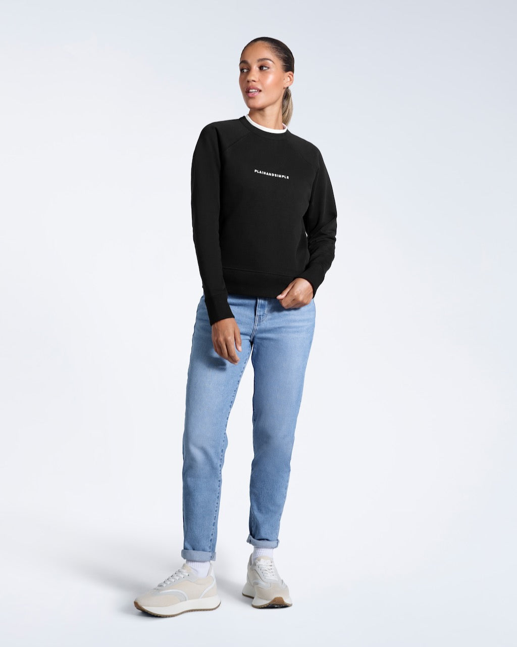 A full body photo of a woman looking away from the camera, modelling a branded black heavyweight organic cotton sweatshirt.