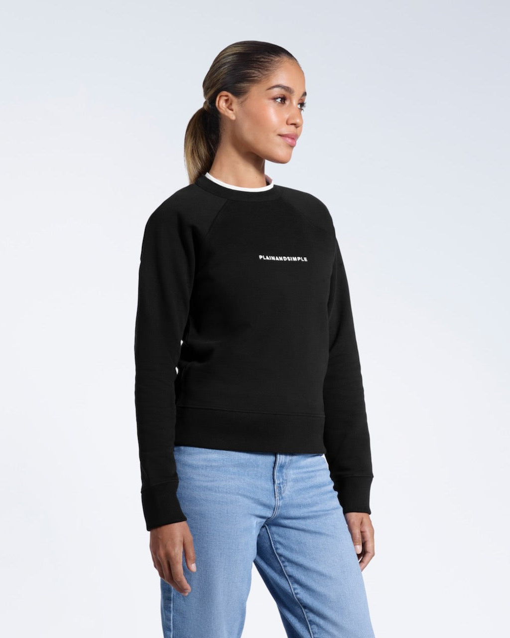 A woman looking away from the camera with her arms by her sides, modelling a branded black heavyweight organic cotton sweatshirt.