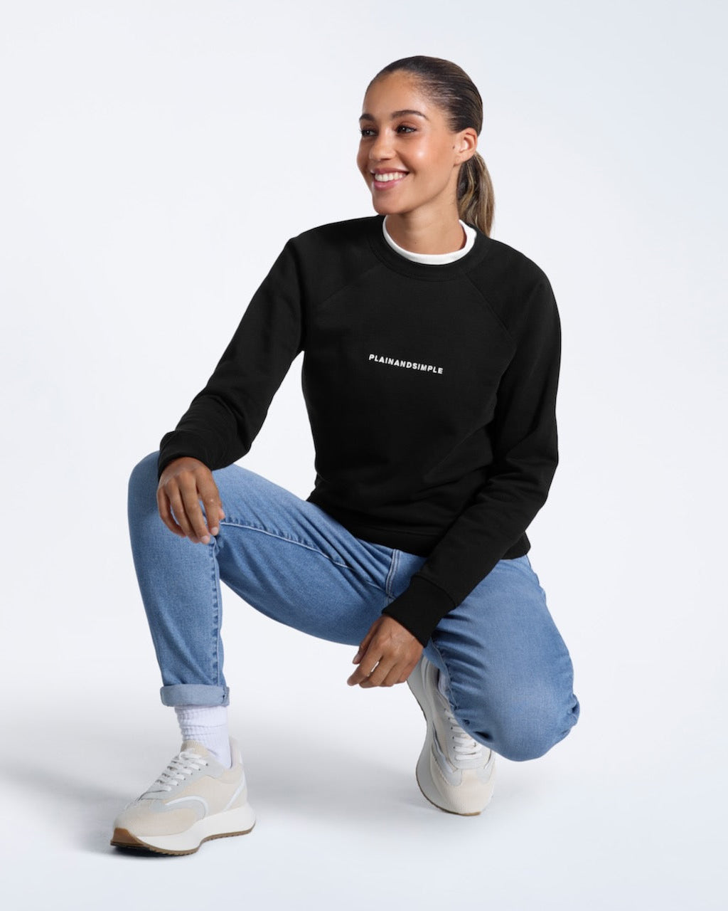 A woman crouching down smiling, modelling a black branded heavyweight organic cotton sweatshirt.