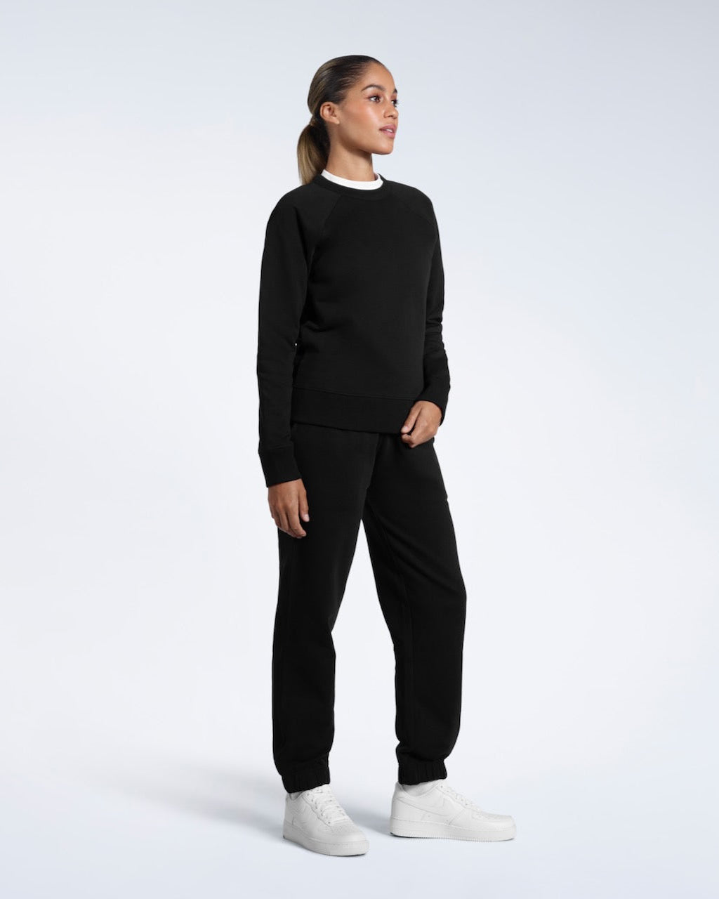 A full body photo of a woman modelling a branded black heavyweight organic cotton sweatshirt with matching joggers.