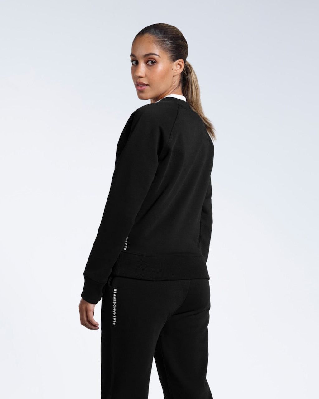 A woman turned to look at the camera, showing the back of her black heavyweight organic cotton sweatshirt with matching joggers.