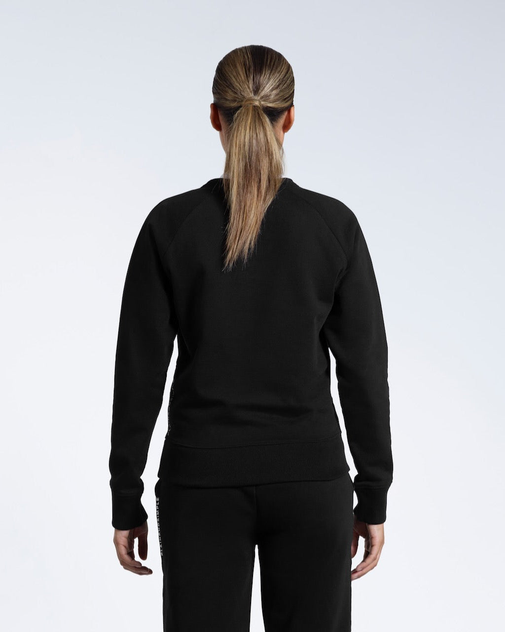 A woman facing away from the camera, showing the back of her black heavyweight organic cotton sweatshirt with matching joggers.