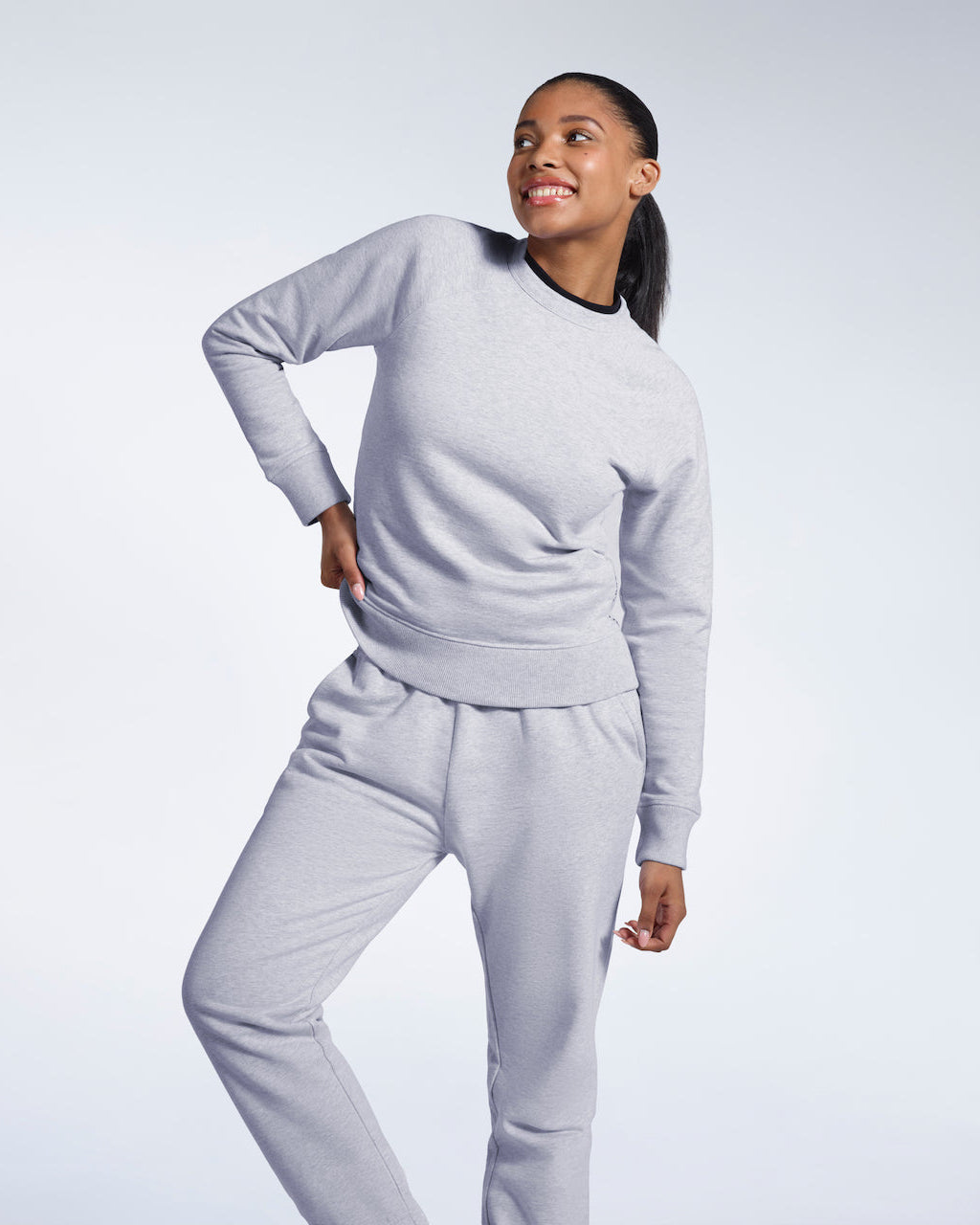 A woman smiling with her hand by her hip, modelling a grey heavyweight organic cotton sweatshirt with matching joggers.