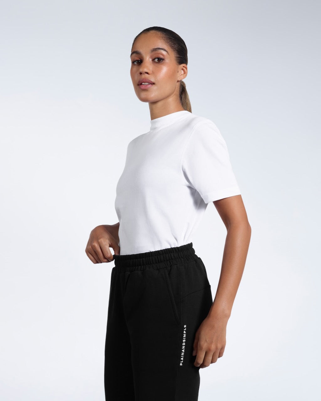 A woman looking into the camera touching her waist, modelling a pair of black heavyweight organic cotton joggers with a white t shirt.