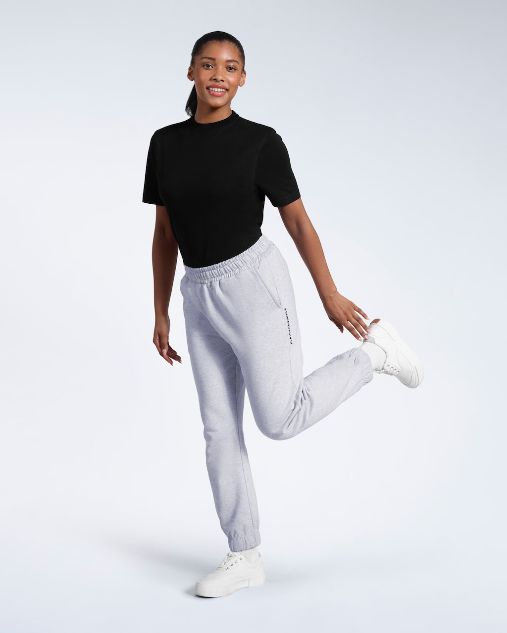 A woman smiling into the camera touching her shoe, modelling a pair of grey heavyweight organic cotton joggers.