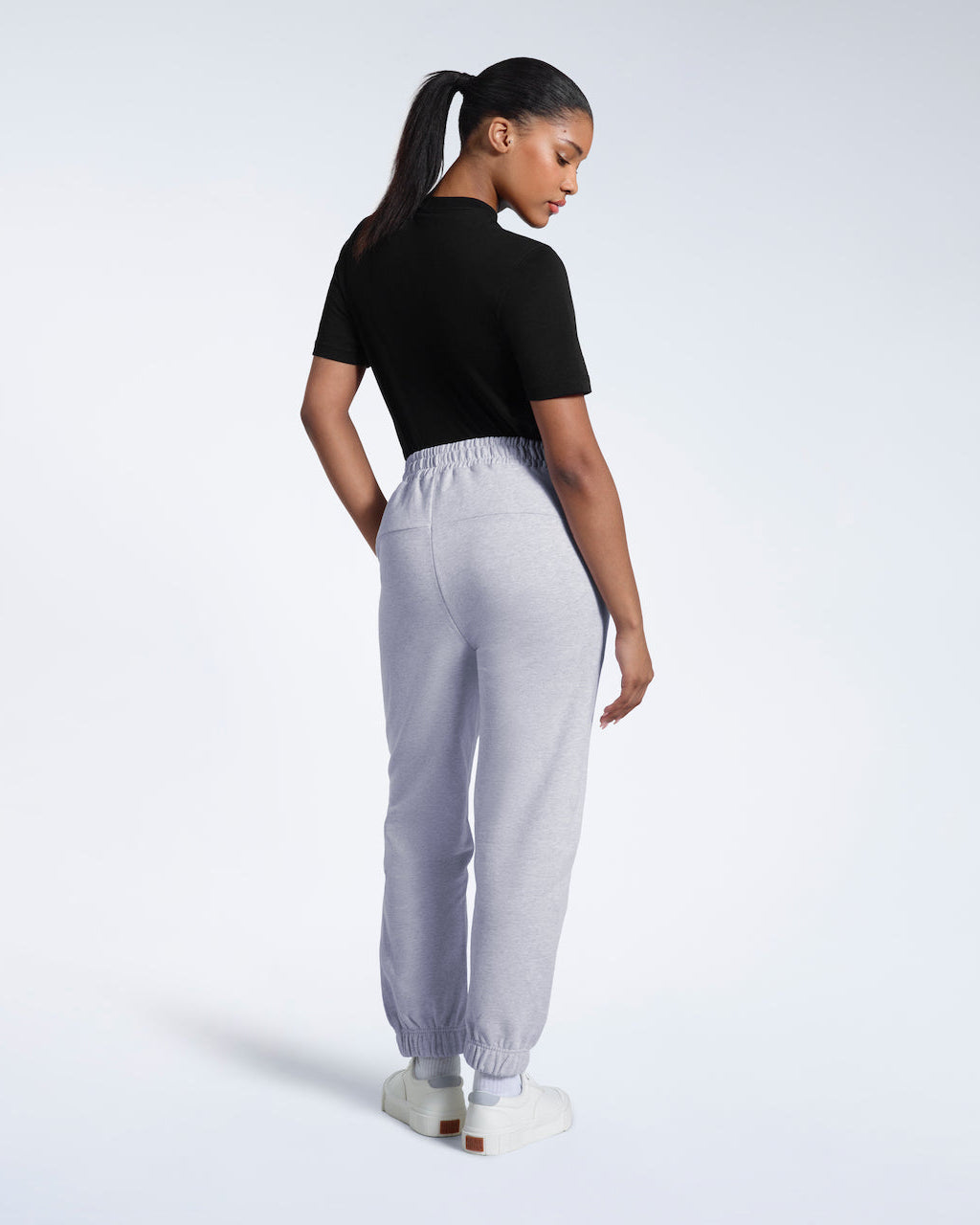 A woman with her back to the camera with one hand in her pocket, modelling a pair of grey heavyweight organic cotton joggers.