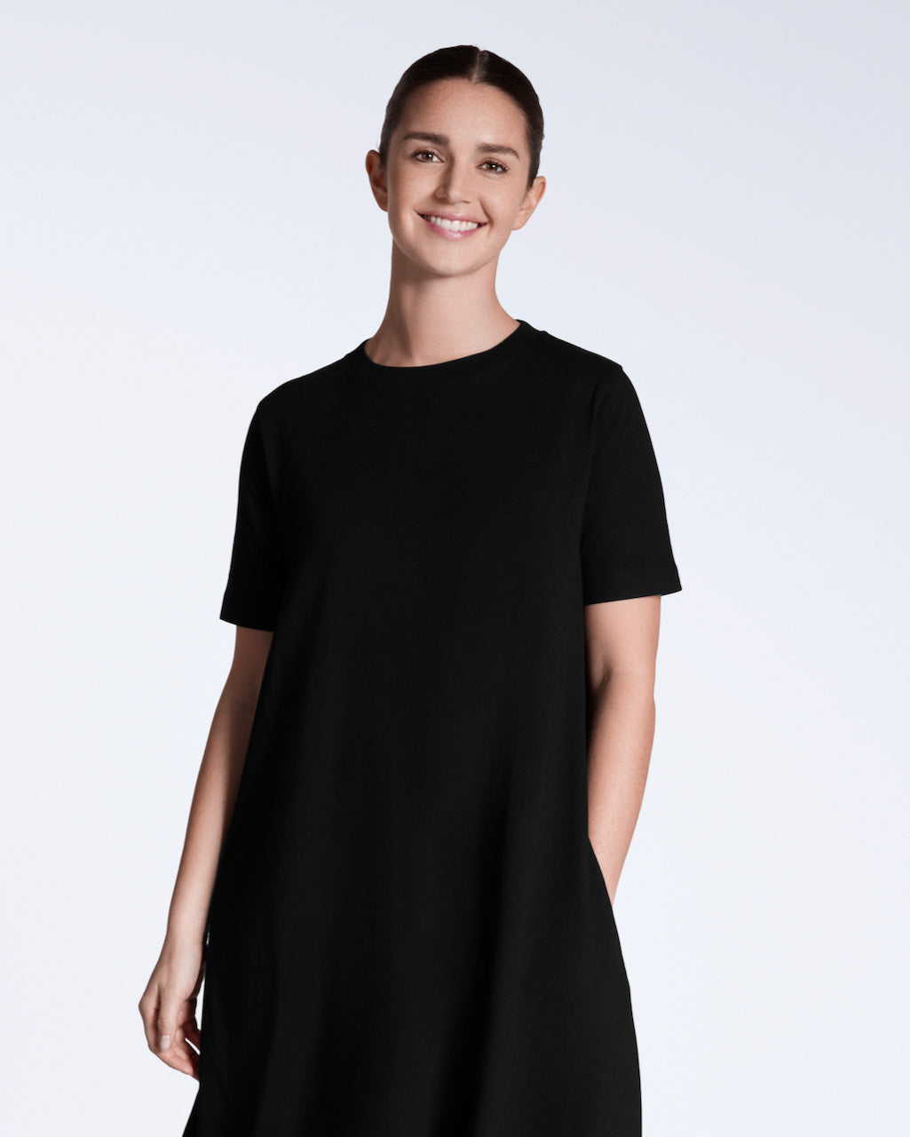 A woman smiling into the camera with her hand in her pocket, modelling a plain black, long line, maxi organic cotton t shirt dress.