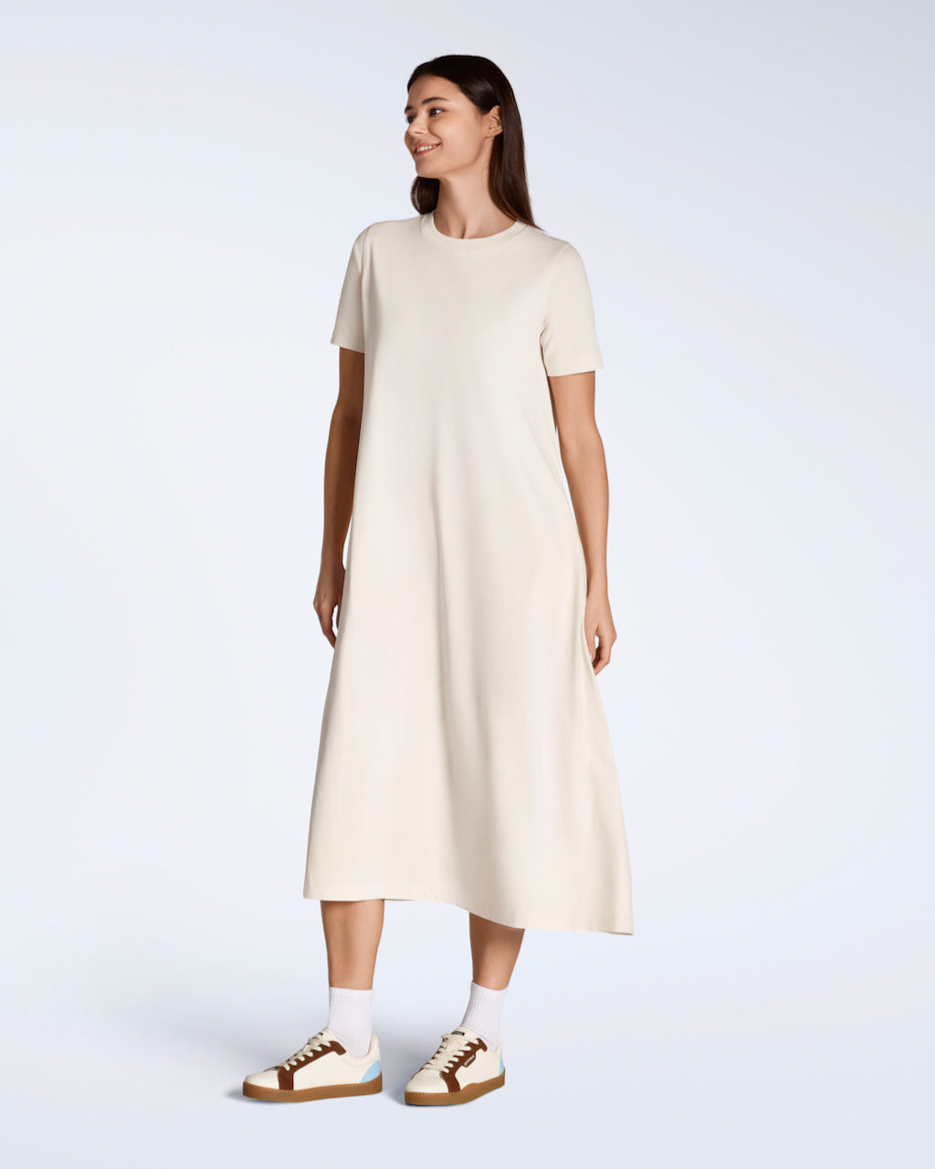 A woman looking away from the camera smiling with her arms by her side, modelling a plain natural coloured, long line, maxi organic cotton t shirt dress.